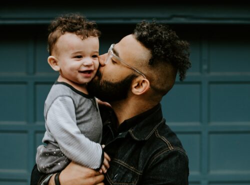 A man holds and kisses his son in front of their home.